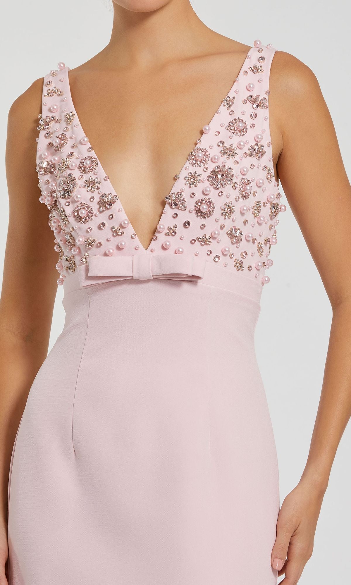 A woman wears the Mac Duggal 2278 beaded-bodice long prom dress, featuring an elegant light pink empire waist, deep V-neck, pearl and bead embellishments, and a small bow at the waist.